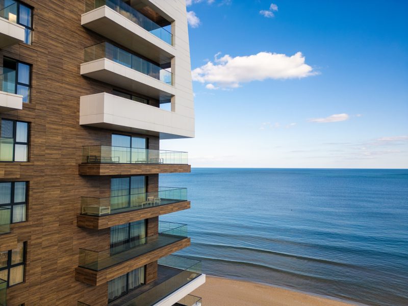 A modern beachfront apartment building with glass balconies overlooks a calm sea. The blue sky with scattered clouds adds to the serene coastal setting.